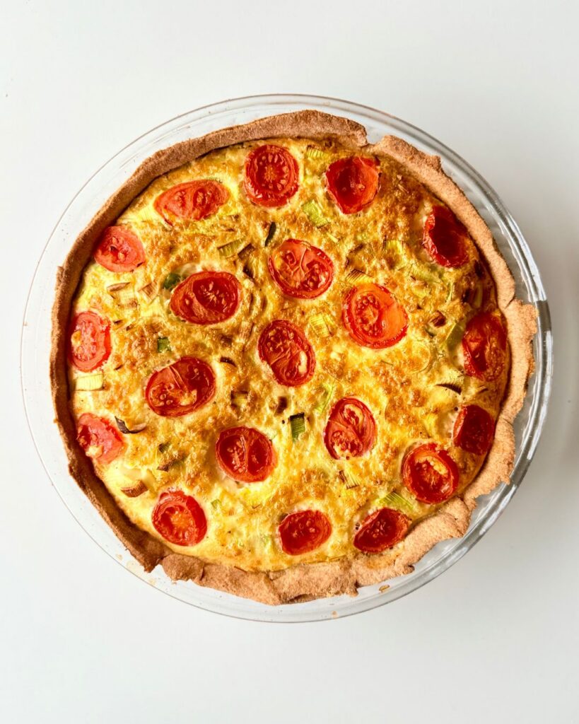 Teljes kiőrlésű tésztából készült paradicsomos-póréhagymás quiche aranybarnára sütve, paradicsommal a tetején.