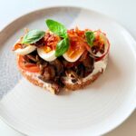 Teljes kiőrlésű kovászos kenyéren ricotta, pirított kakukkfüves gomba, főtt tojás, prosciutto, paradicsom és friss bazsalikom.
