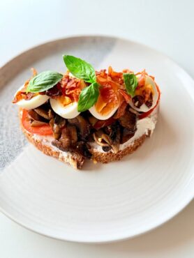 Teljes kiőrlésű kovászos kenyéren ricotta, pirított kakukkfüves gomba, főtt tojás, prosciutto, paradicsom és friss bazsalikom.