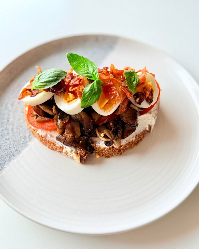 Teljes kiőrlésű kovászos kenyéren ricotta, pirított kakukkfüves gomba, főtt tojás, prosciutto, paradicsom és friss bazsalikom.